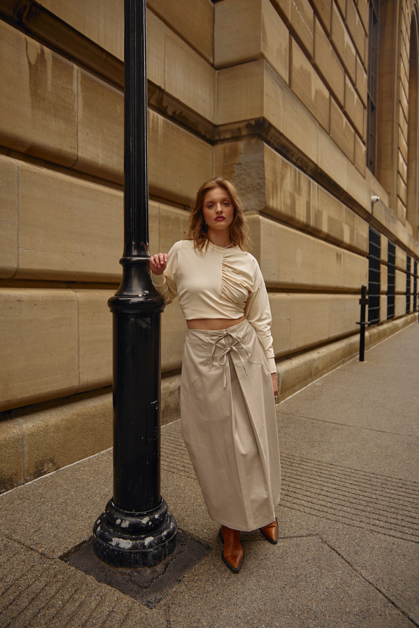 Full front view, female model, cream long sleeve Astrid t-shirt styled to be cropped, Midi length pull-on skirt with front tie closure, slight parachute silhouette, side seam pockets, centre front pleat detail, waist darts, made from a crisp stretch cotton fabric with a slightly peached finish that's soft to the touch. Model is wearing camel coloured pointy leather boots