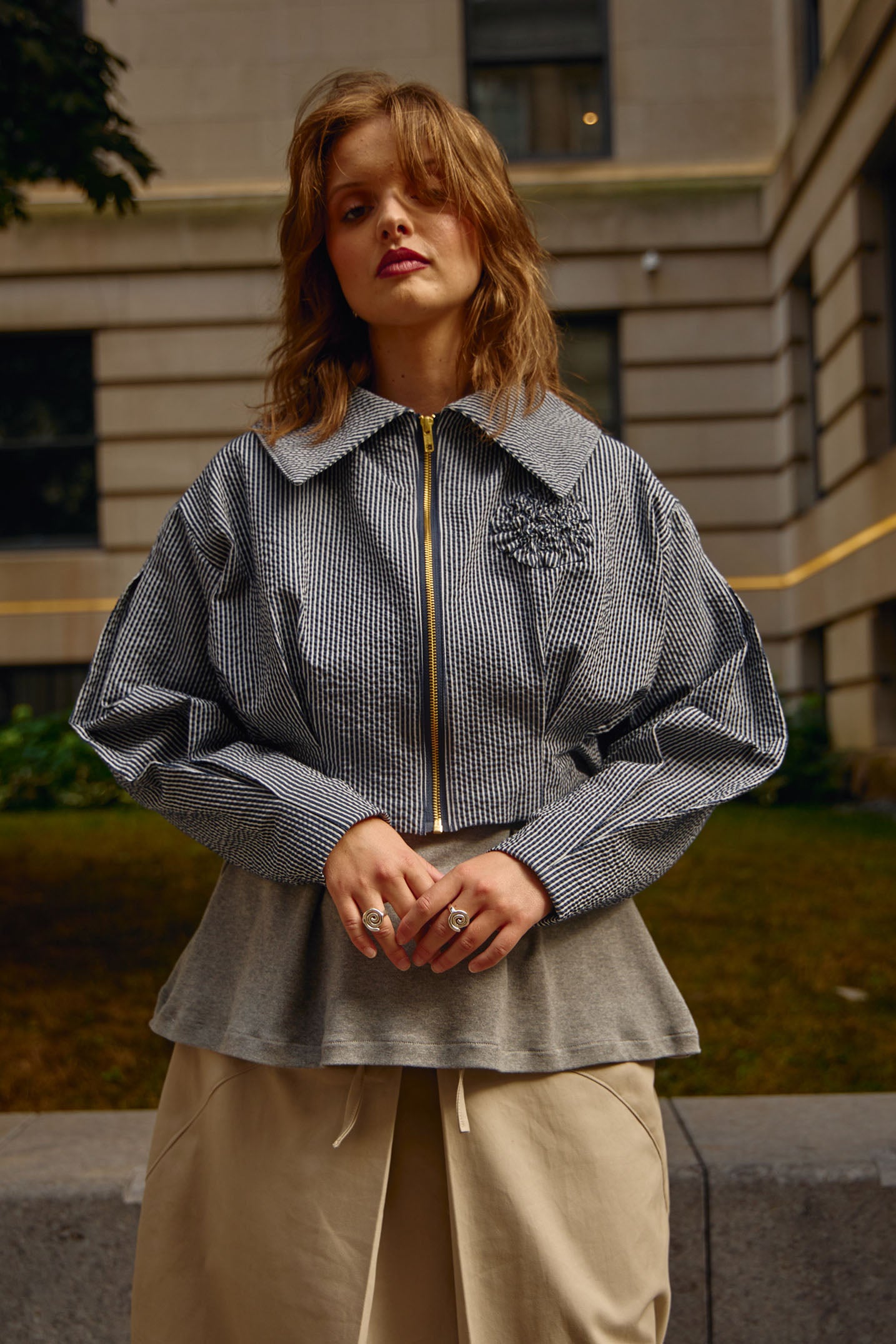 Cropped front view editorial photo of waist-length jacket with matte gold exposed zipper closure, oversized collar, floral appliqué detail at chest and box pleat detailing at waist and sleeves.
Jacket is fully lined with an inner patch pocket.
Made from a beautiful classic navy and white pinstripe seersucker fabric
Styled with a hip length knit heather grey t-shirt and classic khaki colour midi skirt. Model is wearing silver spiral rings on both hands