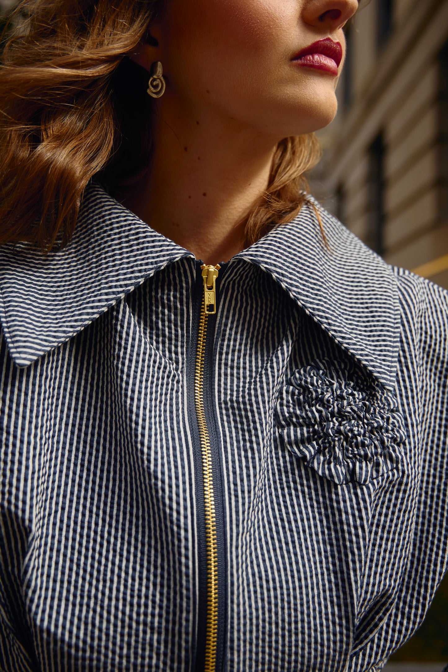 Cropped editorial photo of waist-length jacket with matte gold exposed zipper closure, oversized collar, floral appliqué detail at chest and box pleat detailing at waist and sleeves.
Jacket is fully lined with an inner patch pocket.
Made from a beautiful classic navy and white pinstripe seersucker fabric