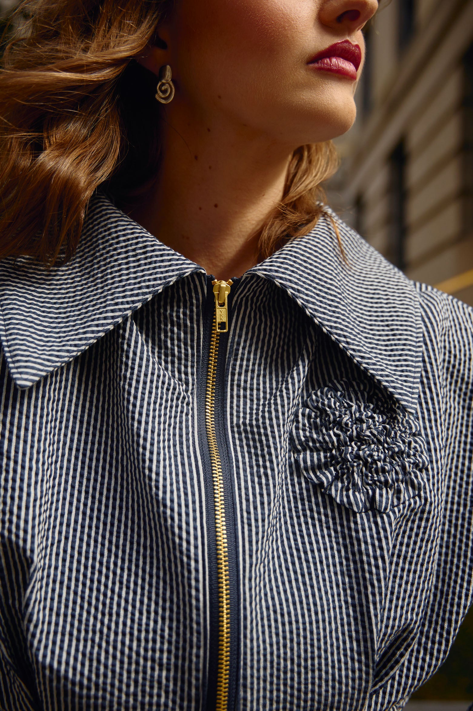 Cropped editorial photo of waist-length jacket with matte gold exposed zipper closure, oversized collar, floral appliqué detail at chest and box pleat detailing at waist and sleeves.
Jacket is fully lined with an inner patch pocket. 
Made from a beautiful classic navy and white pinstripe seersucker fabric