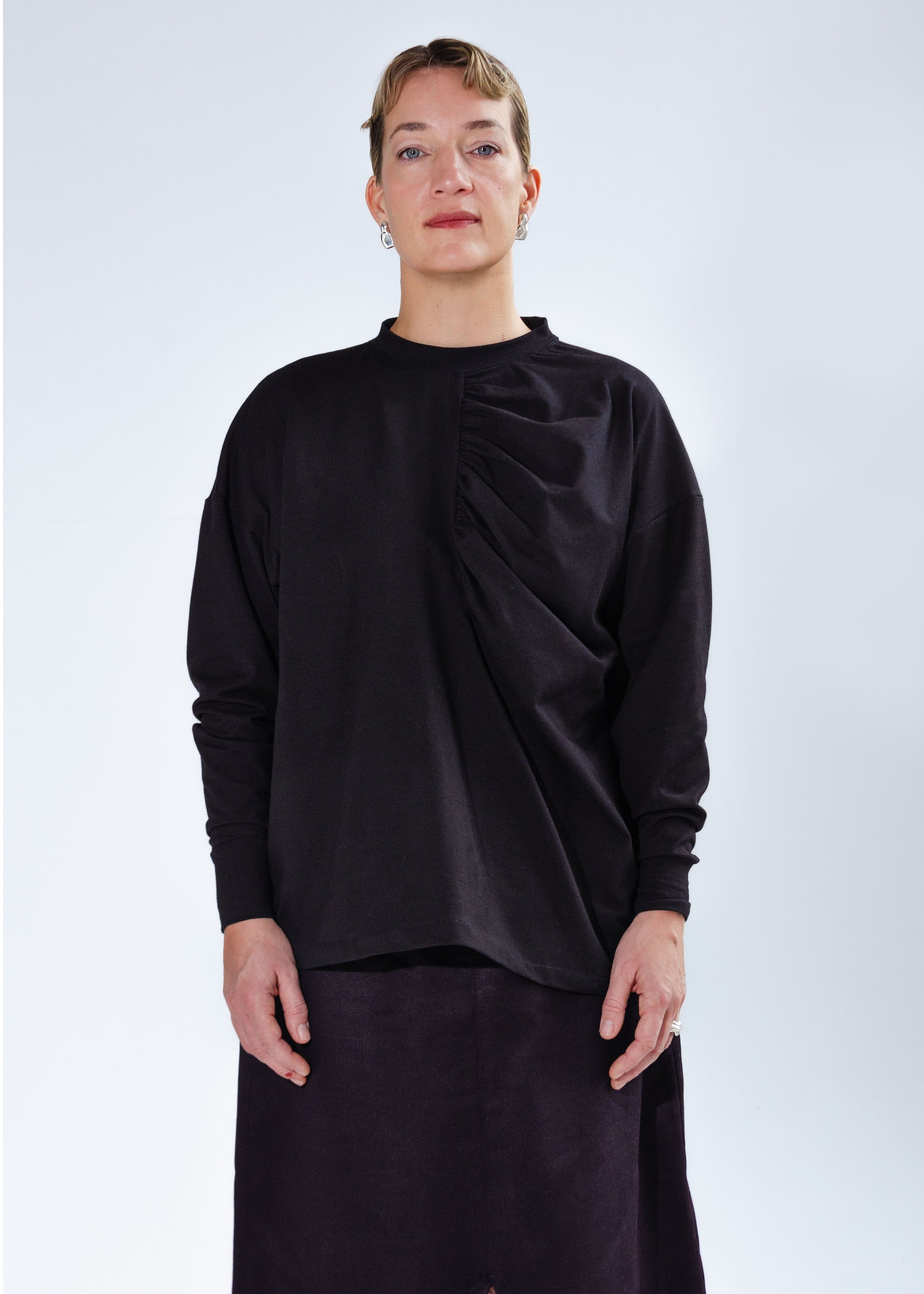 Front view on female model, Oversized long sleeve knit shirt with dropped shoulders and gathering detail at centre front. Black stretchy cotton spandex jersey knit fabric, high neck, neckband, folded cuffs