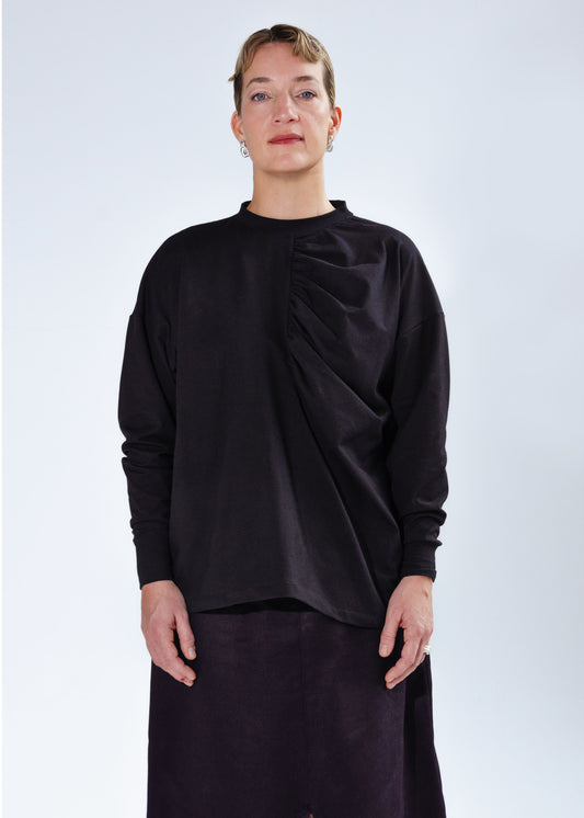 Front view on female model, Oversized long sleeve knit shirt with dropped shoulders and gathering detail at centre front. Black stretchy cotton spandex jersey knit fabric, high neck, neckband, folded cuffs