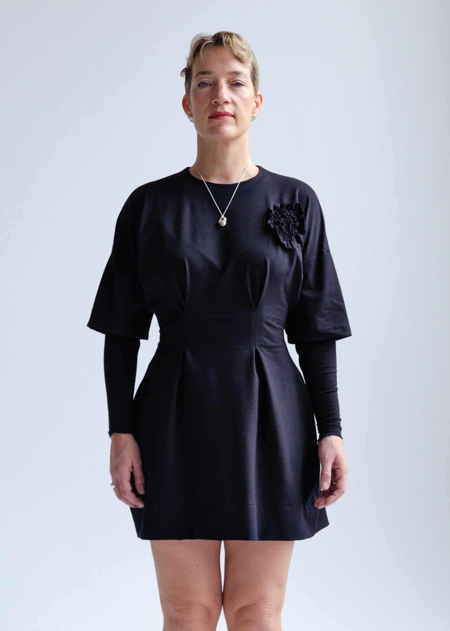 Front view, female model, drop shoulder, black knit mini dress with fit flare waist, double layer sleeve, appliqué flower detail at chest.
Cozy, stretchy cotton spandex knit jersey fabric.Our version of the "Little Black Dress"