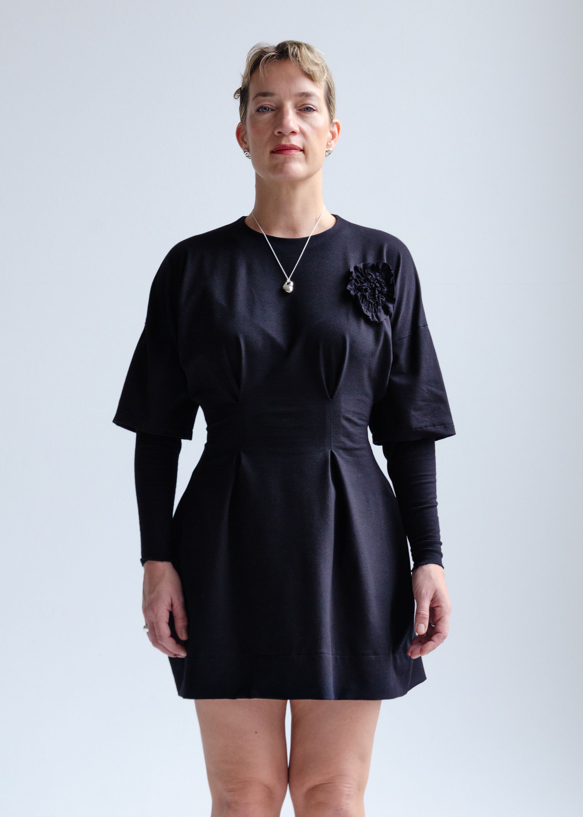 Front view, female model, drop shoulder, black knit mini dress with fit flare waist, double layer sleeve, appliqué flower detail at chest.
Cozy, stretchy  cotton spandex knit jersey fabric.Our version of the "Little Black Dress"