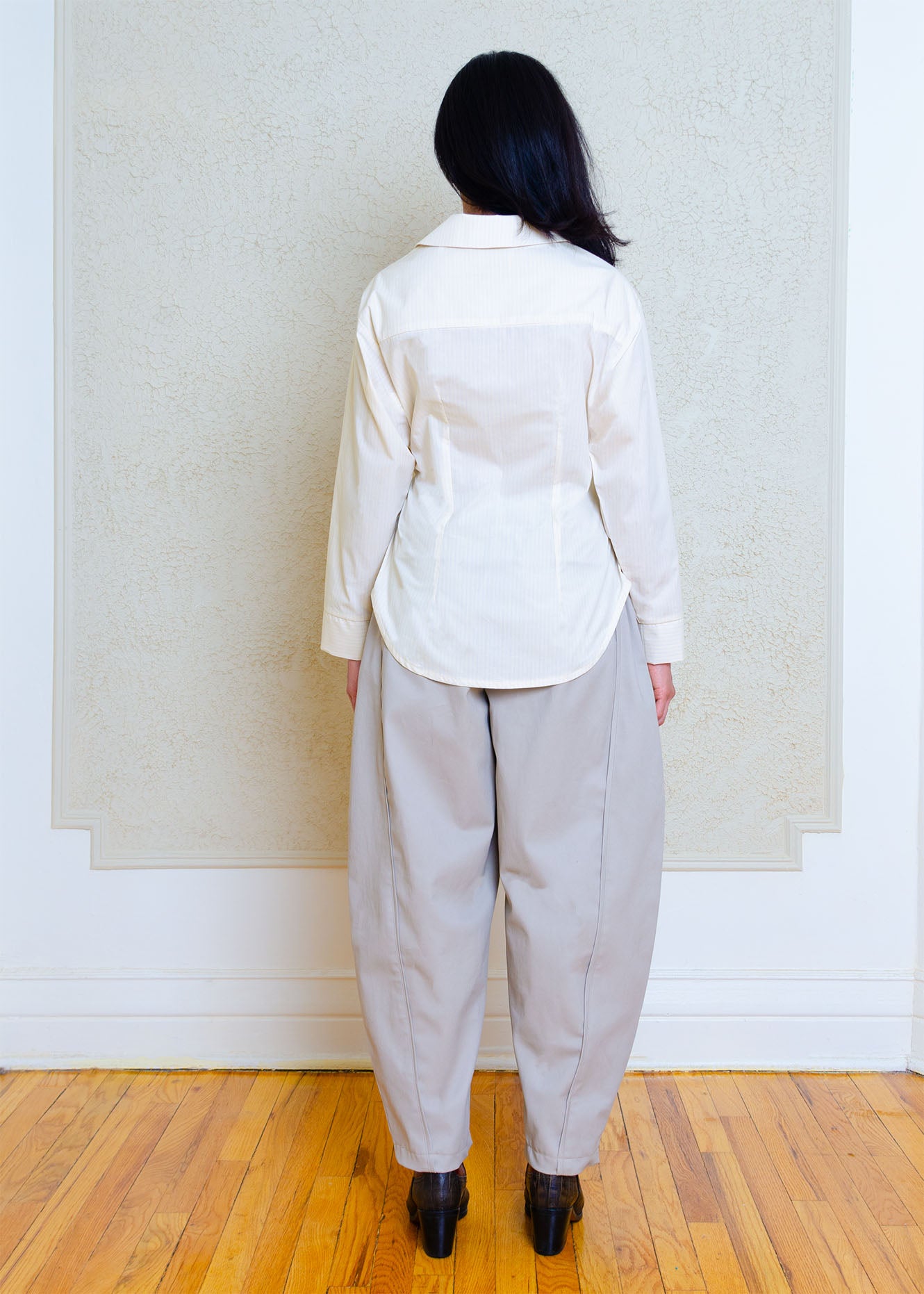 Back view. Cotton poplin shirt with asymmetrical front, adjustable waist closure with matte tortoiseshell print slider, gathering at waist side seams, shirt collar & patch pocket.Featuring an exaggerated baseball hem & back darts for a flattering, slightly fitted look.
Lightweight light yellow cotton poplin fabric features a subtle menswear-influenced stripe.