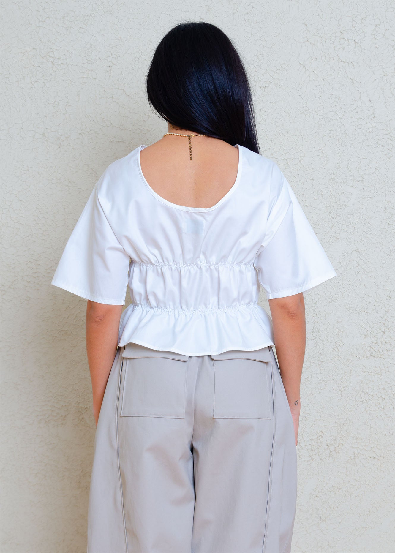 Back view. Rouched short sleeve top with dropped shoulders and low back. Elasticized waist detailing allows for a greater size variation. Crisp cotton poplin fabric