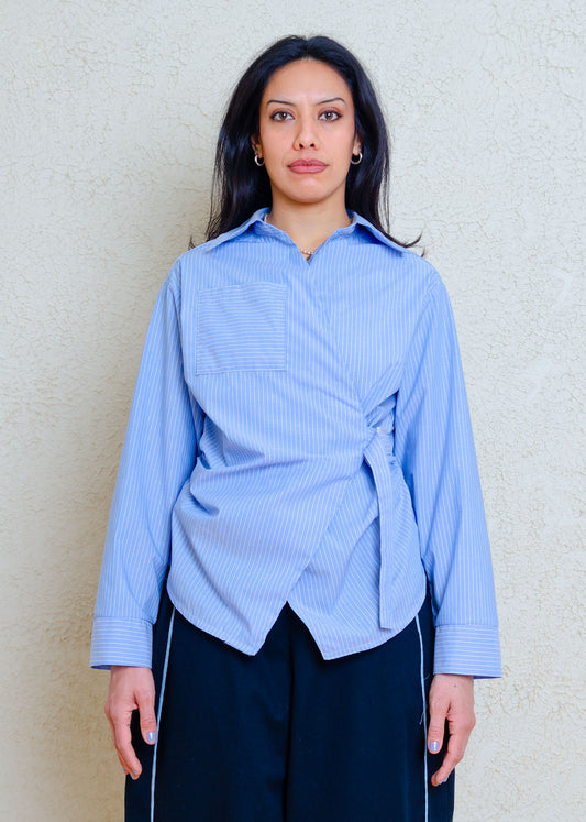Cropped  full view on model. Cotton poplin shirt with asymmetrical front, adjustable waist closure with matte tortoiseshell print slider, gathering at waist side seams, shirt collar & patch pocket.Featuring an exaggerated baseball hem & back darts for a flattering, slightly fitted look.
Lightweight cotton poplin fabric features a subtle menswear-influenced stripe 
Styled with black barrel pants with light denim trim