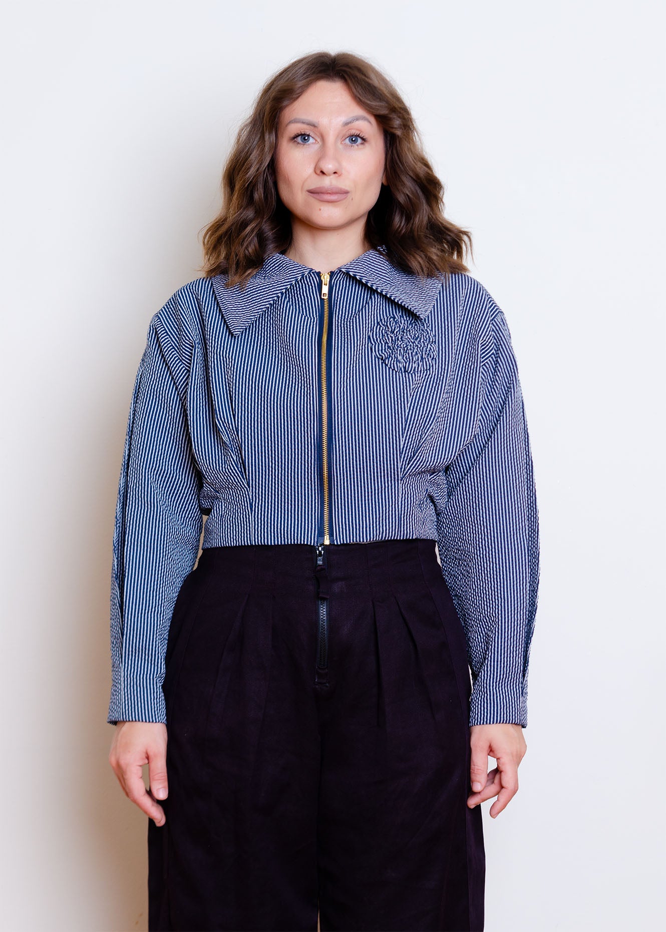Cropped front image on female model. Waist-length jacket with exposed zipper closure, oversized collar, floral appliqué detail at chest and box pleat detailing at waist and sleeves.
Jacket is fully lined with an inner patch pocket.
Made from a beautiful classic navy and white pinstripe seersucker fabric