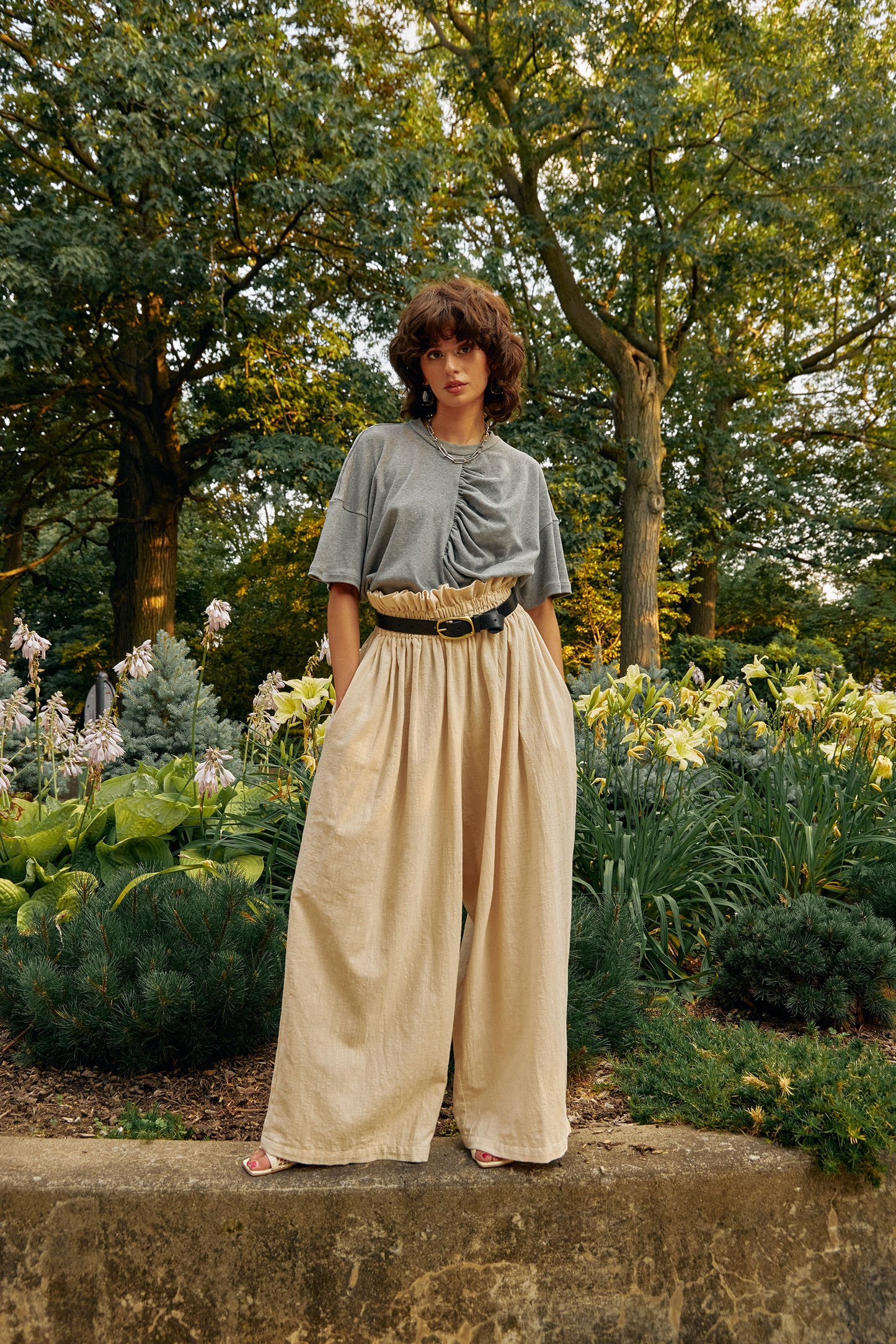 Oversized t-shirt with dropped shoulders and centre front gathering detail made from soft organic cotton.Interlock knit fabric, medium heather grey colour. A classic, elevated basic.
Editorial photo in a park during Spring. Styled with cream high waisted, very wide leg pants. Model has her hands in the side seam pockets.