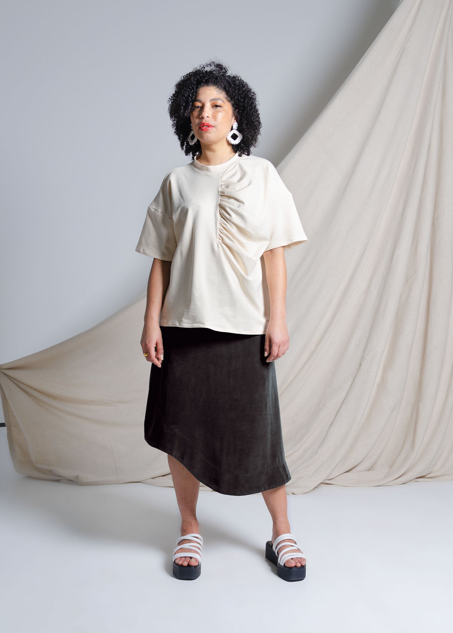 Curly haired model wears oversize cream cotton tank top with dropped shoulders and gathering detail at front. Black asymmetrical hem midi skirt made from textured cupro blend fabric