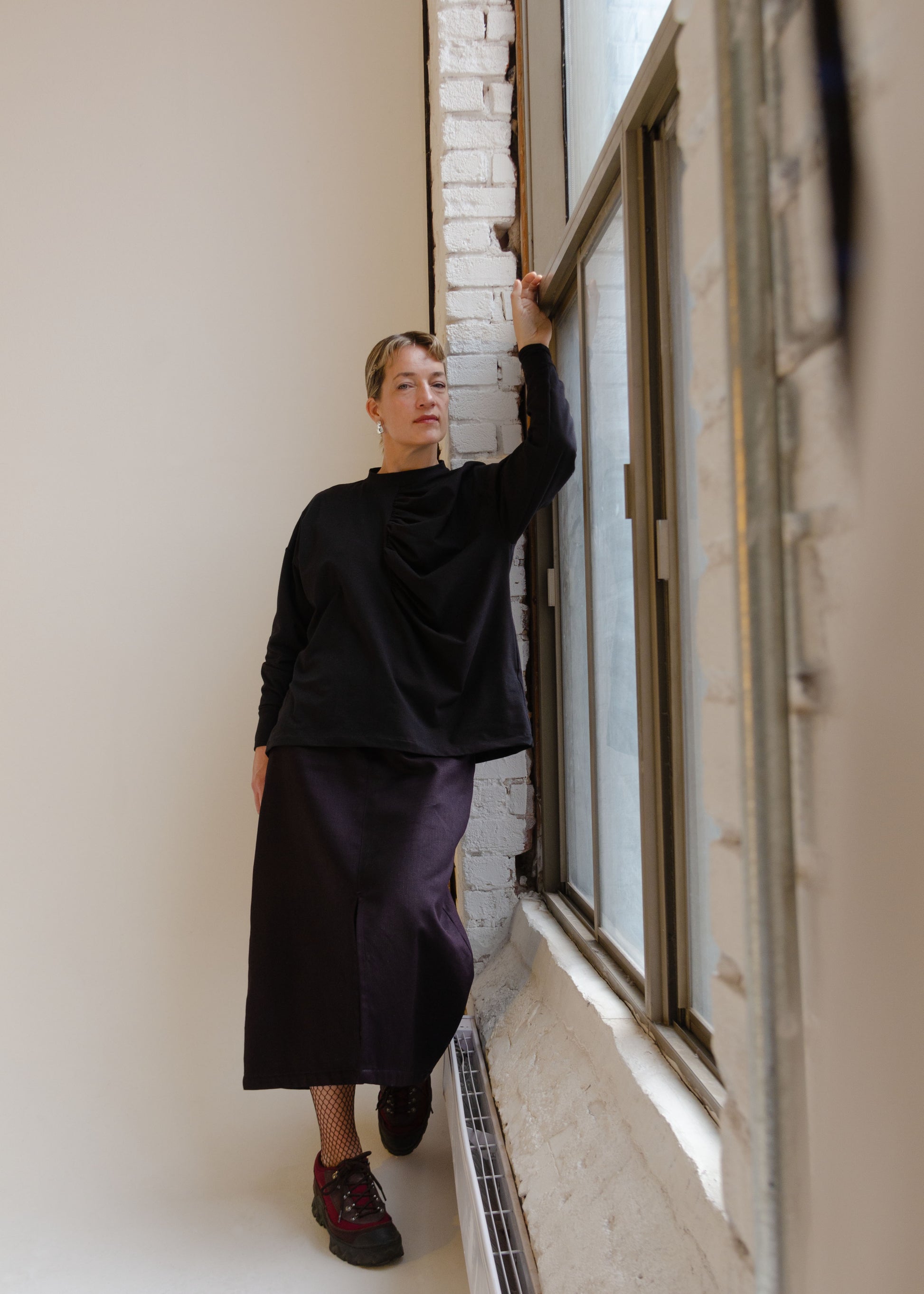 Full Front view on female model wearing midi length denim skirt, fishnet tights and burgundy running shoes., Oversized long sleeve knit shirt with dropped shoulders and gathering detail at centre front. Black stretchy cotton spandex jersey knit fabric, high neck, neckband, folded cuffs 