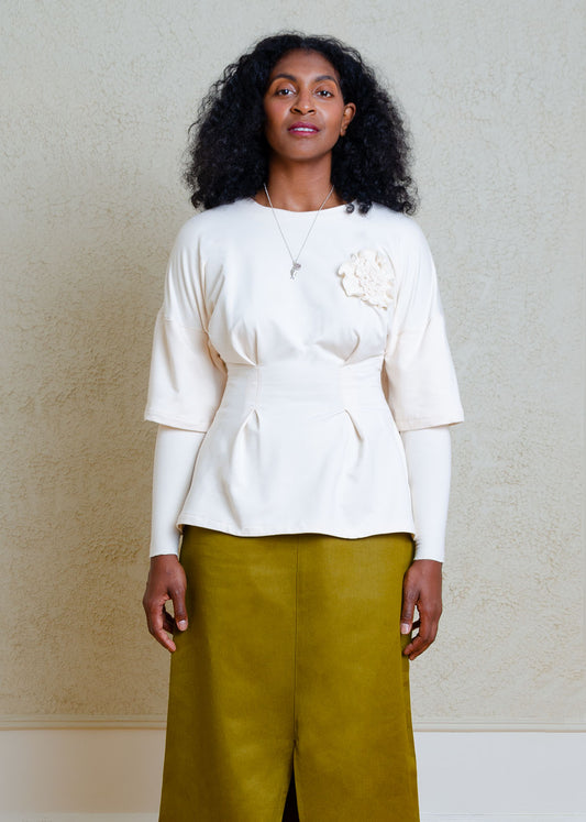 Woman wearing a cream long sleeve top with double layer sleeves and floral appliqué detail at the chest and green skirt against a plain background