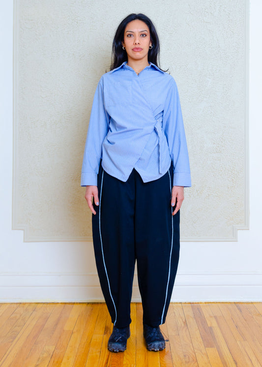 Front full view on model. Cotton poplin shirt with asymmetrical front, adjustable waist closure with matte tortoiseshell print slider, gathering at waist side seams, shirt collar & patch pocket.Featuring an exaggerated baseball hem & back darts for a flattering, slightly fitted look.

Lightweight cotton poplin fabric features a subtle menswear-influenced stripe 

Styled with black barrel pants with light denim trim and black running shoes 