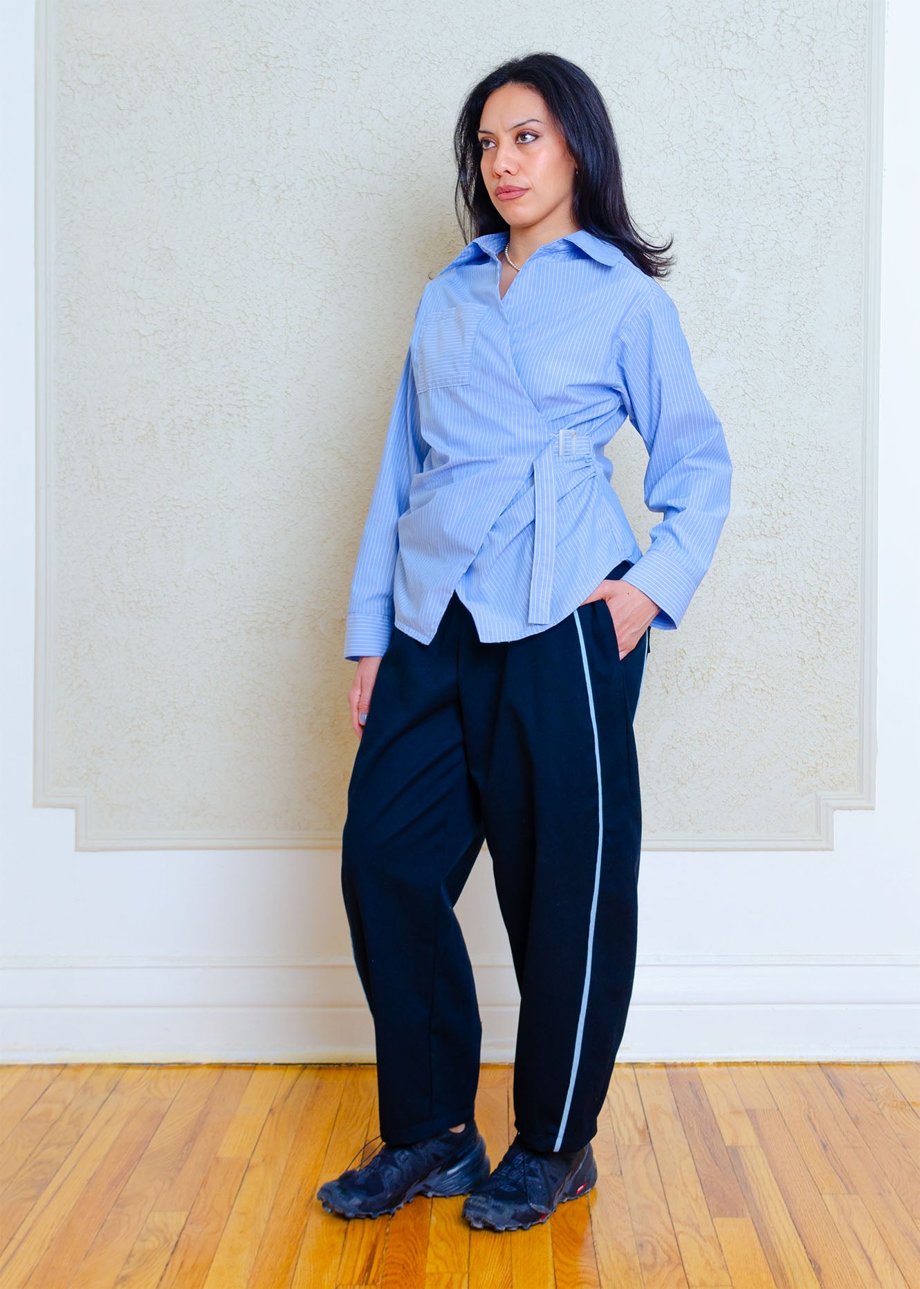 There quarter  full view on model. Cotton poplin shirt with asymmetrical front, adjustable waist closure with matte tortoiseshell print slider, gathering at waist side seams, shirt collar & patch pocket.Featuring an exaggerated baseball hem & back darts for a flattering, slightly fitted look.

Lightweight cotton poplin fabric features a subtle menswear-influenced stripe 

Styled with black barrel pants with light denim trim and black running shoes 