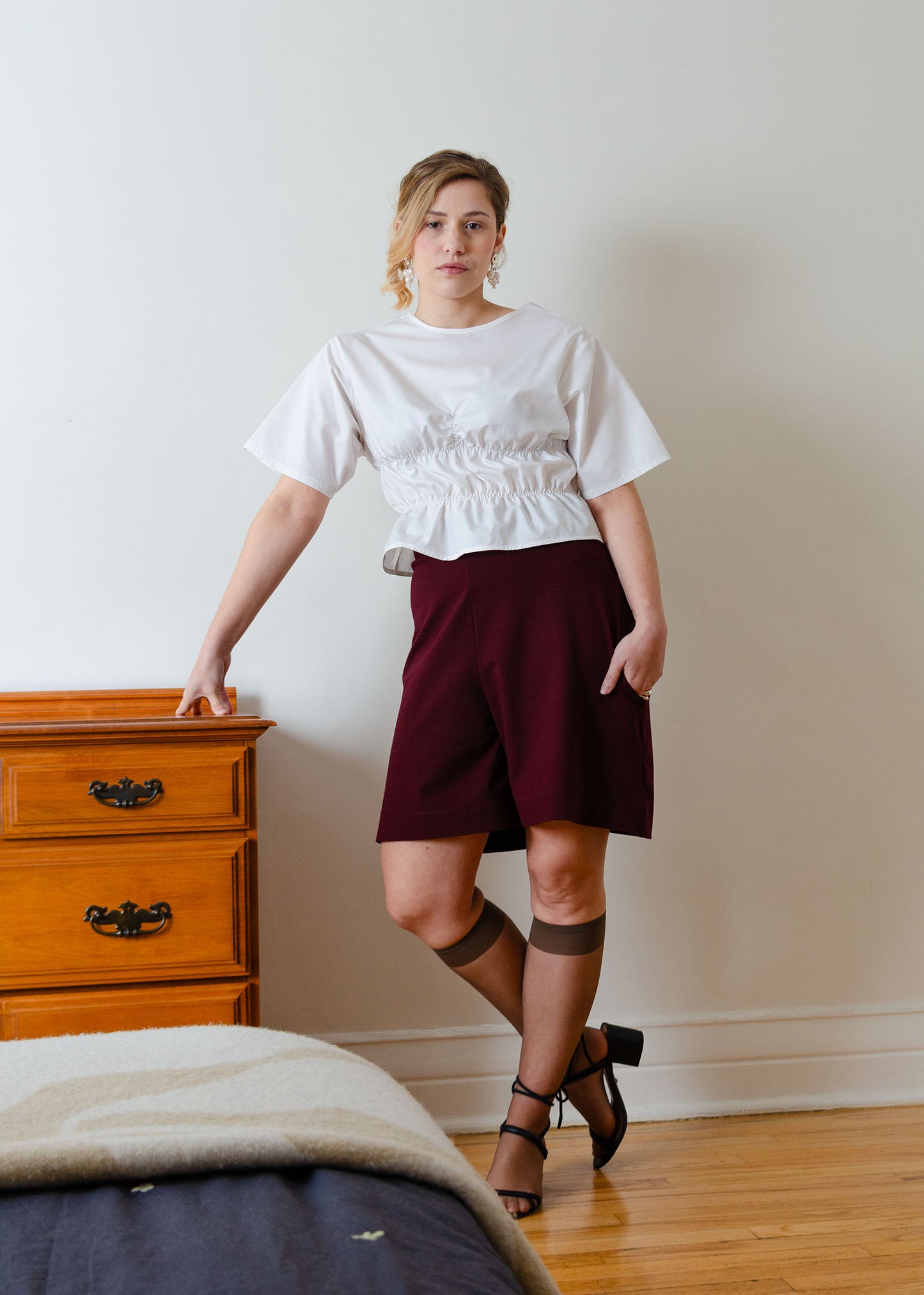 Campaign shoot, blonde model in bedroom posing in front of the mirror. Wearing white cotton poplin t-shirt with rouching, pull on chocolate brown knit jorts, knee high brown sheer stockings and black lace up sandals 