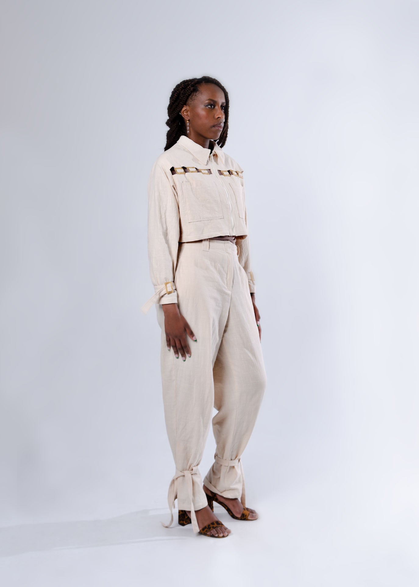 Model wears matching cream outfit. Long sleeve cropped jacket with centre front exposed zipper, chest pockets and cutout details. High waisted pants, tied at the ankles